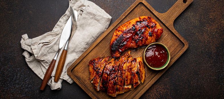 Grilled turkey or chicken marinated fillet with red sauce served and sliced on wooden cutting board on stone brown background from above, poultry breast barbecue