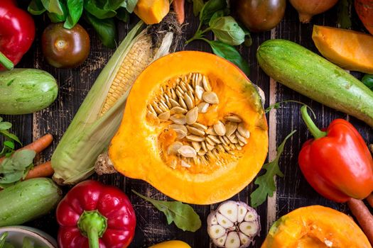 Fresh vegetables scattered on a old rustic dark textured table. Autumn background. Healthy eating. Sliced pumpkin, zucchini, squash, bell peppers, carrots, onions, cut garlic, tomatoes, eggplant, corn cob, rucola and basil. Top view