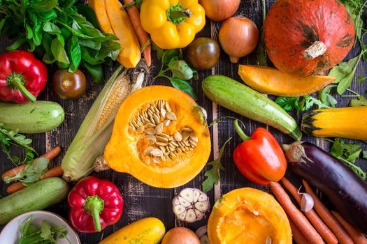 Fresh vegetables scattered on a old rustic dark textured table. Autumn background. Healthy eating. Sliced pumpkin, zucchini, squash, bell peppers, carrots, onions, cut garlic, tomatoes, eggplant, corn cob, rucola and basil. Top view