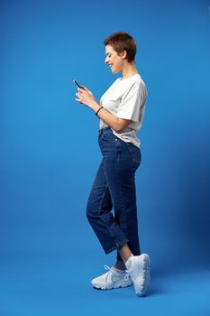 Cheerful attractive young woman holding smartphone, enjoying chatting or online shopping against blue background, close up