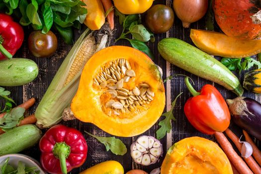 Fresh vegetables scattered on a old rustic dark textured table. Autumn background. Healthy eating. Sliced pumpkin, zucchini, squash, bell peppers, carrots, onions, cut garlic, tomatoes, eggplant, corn cob, rucola and basil. Top view