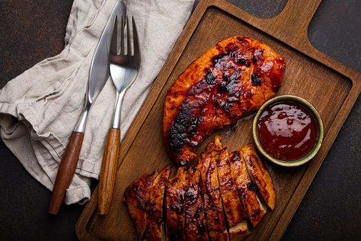 Grilled turkey or chicken marinated fillet with red sauce served and sliced on wooden cutting board on stone brown background from above, poultry breast barbecue