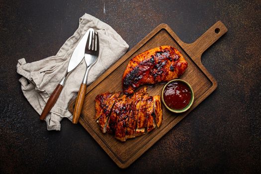 Grilled turkey or chicken marinated fillet with red sauce served and sliced on wooden cutting board on stone brown background from above, poultry breast barbecue