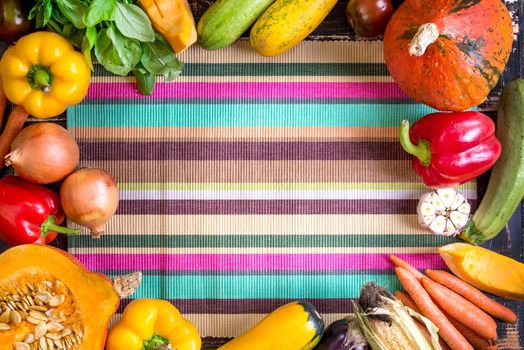Fresh vegetables on a colorful striped kitchen towel and old rustic dark textured table. Autumn background. Healthy eating frame. Sliced pumpkin, bell peppers, carrots, onions, cut garlic, tomatoes, rucola and basil. Top view. Space for text