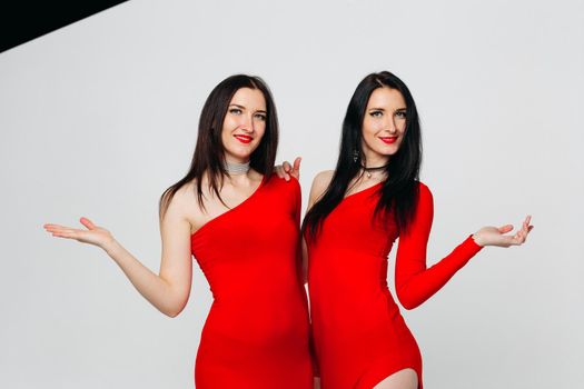 Two sexy and beautiful sisters twins in red dresses posing, looking at camera holding hands up. Pretty dancing ladies with long hair. Fashionable and stylish girls. Shopping, fashion, party.