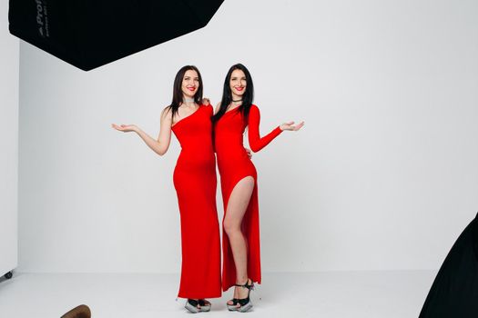 Two sexy and beautiful sisters twins in red dresses posing, looking at camera holding hands up. Pretty dancing ladies with long hair. Fashionable and stylish girls. Shopping, fashion, party.