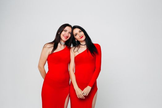 Two sexy and beautiful sisters twins in red dresses posing, looking at camera holding hands up. Pretty dancing ladies with long hair. Fashionable and stylish girls. Shopping, fashion, party.