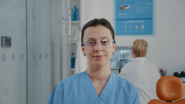 Portrait of orthodontic nurse looking at camera in dentistry office. Woman assistant wearing protective glasses before attending stomatological procedure at oral care clinic. Close up