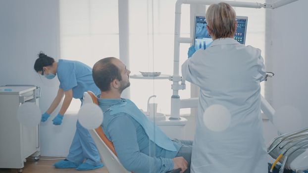 Medic pointing at x ray scan diagnosis on monitor in dentistry cabinet, explaining radiography results to patient in dental chair. Dentist doing checkup examination with man in pain.