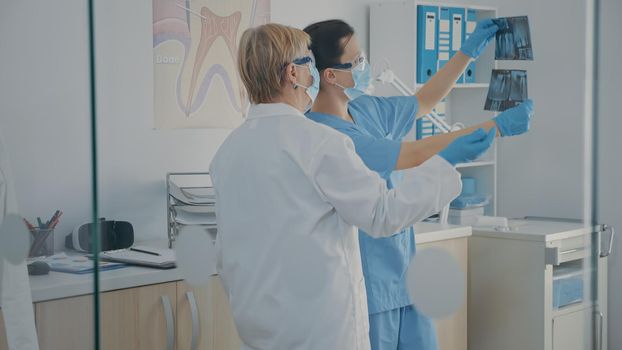 Senior dentist and assistant examining denture x ray results before doing oral care inspection on patient. Team of orthodontists looking at teeth radiography scan in stomatology cabinet.