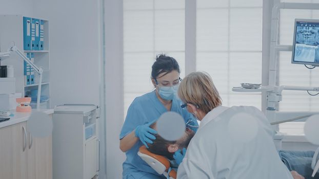 Stomatological team performing drill procedure on patient with toothache, using dental tools and equipment at oral care clinic. Nurse and orthodontist doing teeth inspection to remove caries.