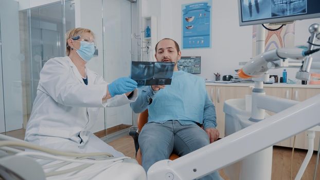 Stomatologist showing denture x ray results to patient at checkup visit, explaining radiography scan diagnosis to understant cavity problems and toothache. Dentist doing oral care consultation.