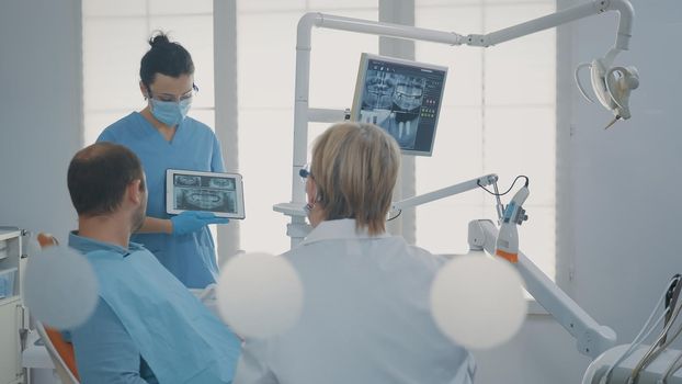 Dentist pointing at denture radiography on digital tablet, explaining diagnosis to patient with caries. Medic showing x ray scan results to man in pain to do surgical procedure in cabinet.