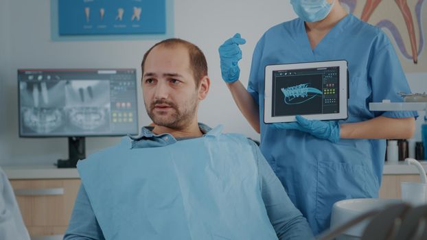 Dentistry assistant showing x ray scan results on tablet to patient, explaining cavity problems and caries. Woman nurse analyzing dentition radiography with man at stomatology consultation.