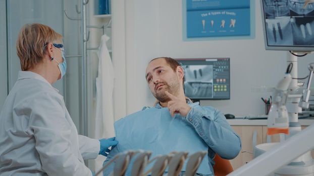 Senior dentist examining patient with serious toothache, getting ready to do stomatological consultation with dental tools in cabinet. Medic treating man in pain with oral care problems.