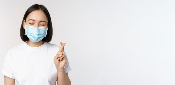 Covid-19, healthcare and medical concept. Image of asian girl in medical face mask, cross fingers, praying, making wish and smiling, standing over white background.