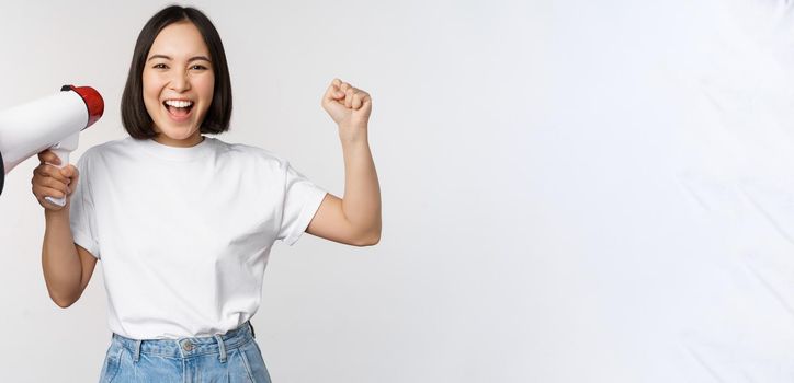 Happy asian woman shouting at megaphone, making announcement, advertising something, standing over white background.