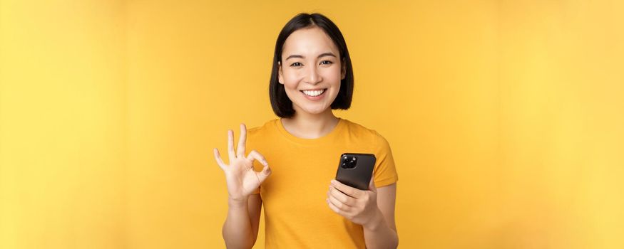 Happy smiling asian girl holding mobile phone and showing okay, recommending application on smartphone, standing over yellow background.