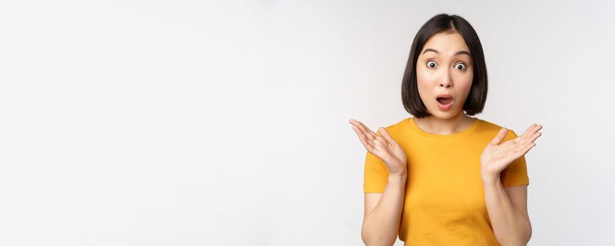 Close up portrait of asian woman looking surprised, wow face, staring impressed at camera, standing over white background in yellow t-shirt.