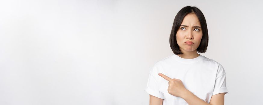 Angry and upset young asian woman pointing left, looking with regret or jealousy, feel unfair, standing over white background. Copy space