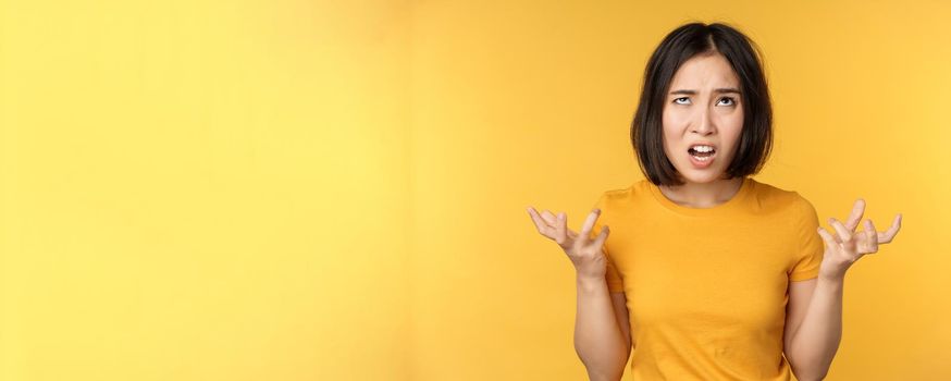 Image of angry asian woman, shouting and cursing, looking outraged, furious face expression, standing over yellow background.