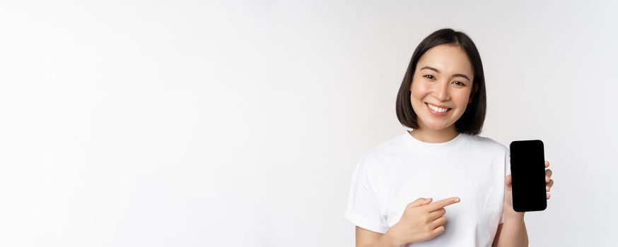Smiling asian woman pointing finger at smartphone screen, showing application interface, mobile phone website, standing over white background.