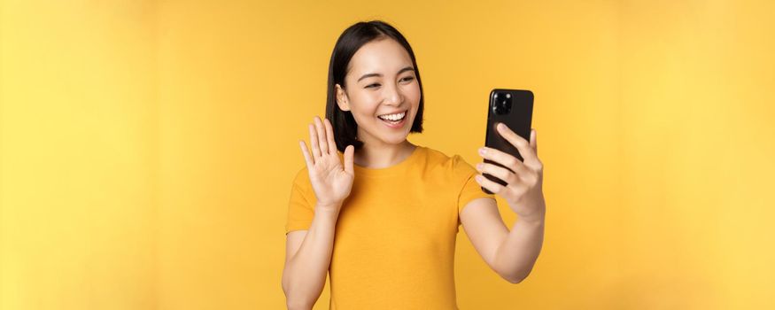 Image of happy, beautiful asian girl video chatting, talking on smartphone application, standing against yellow background. Copy space