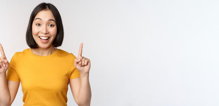 Happy beautiful asian girl showing advertisement, pointing fingers up, standing over white background.