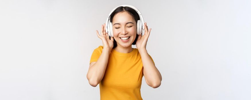 Happy asian girl dancing, listening music on headphones and smiling, standing in yellow tshirt against white background. Copy space