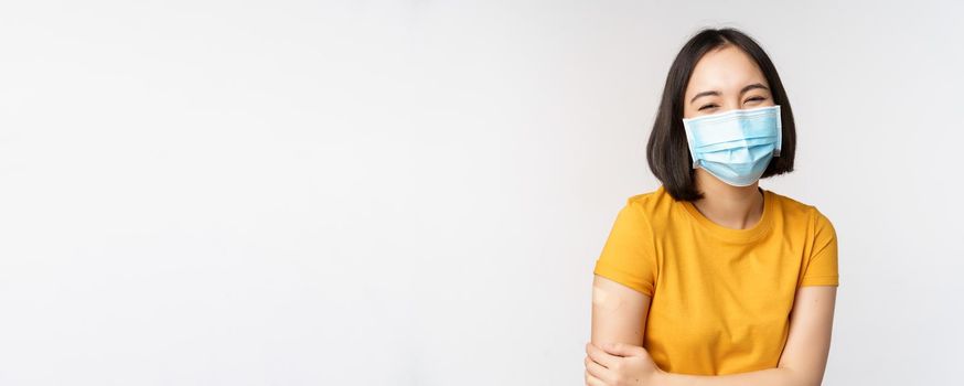 Healthy and happy asian woman in medical face mask, has band aid on shoulder after covid-19 vaccination, got vaccinated from omicron delta variant, whtie background.