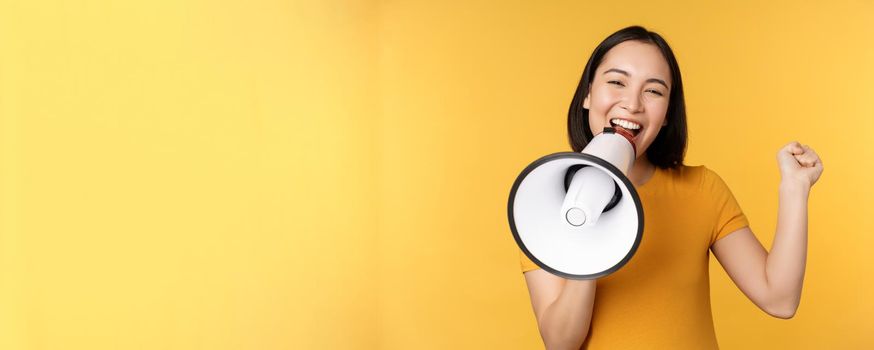 Smiling asian woman standing with megaphone, announcing smth, advertising product, standing over yellow background.