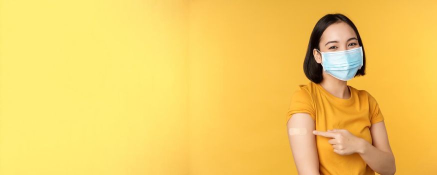 Vaccination and covid-19 pandemic concept. Smiling asian woman in medical face mask, showing her shoulder with band aid after vaccinating from coronavirus, yellow background.