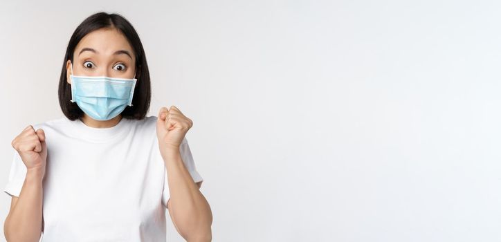 Covid-19, healthcare and medical concept. Enthusiastic asian woman in medical face mask, dancing and celebrating, winning, achieve goal, standing over white background.