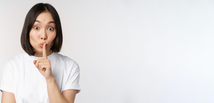 Close up portrait of young beautiful asian girl shushing, has secret, keep quiet silence gesture, press finger to lips, standing in tshirt over white background.