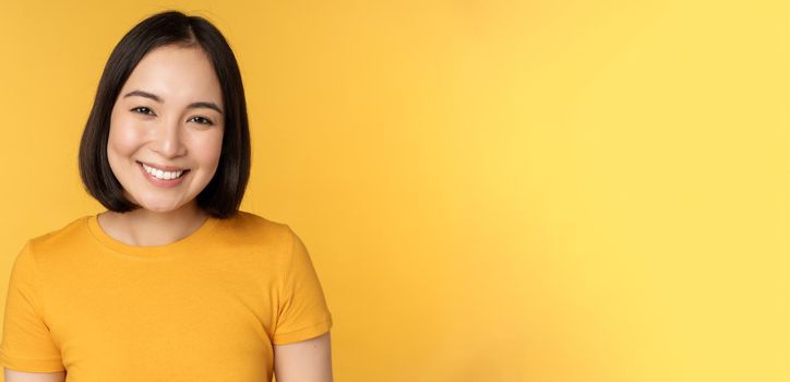 Close up portrait of beautiful asian woman smiling, looking cute and tender, standing against yellow background.