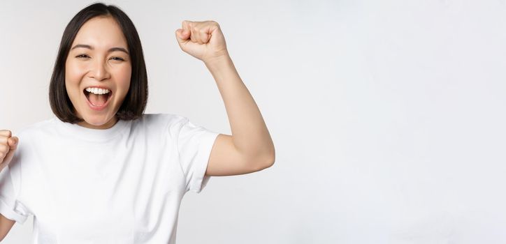 Portrait of enthusiastic asian woman winning, celebrating and triumphing, raising hands up, achieve goal or success, standing over white background.