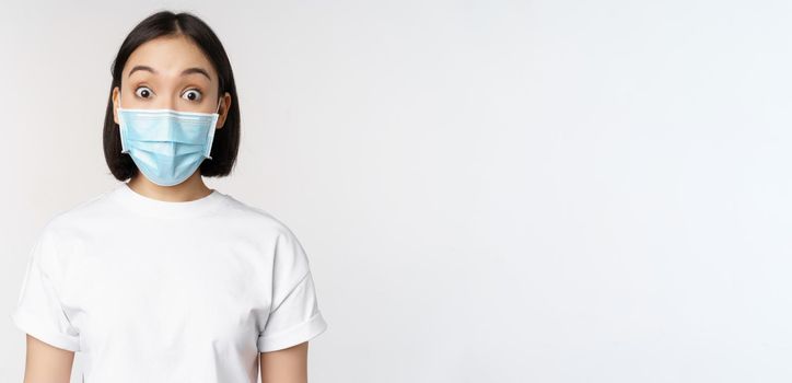 Health and covid pandemic concept. Image of korean woman in face medical mask looking surprised, reacting amazed, standing over white background.