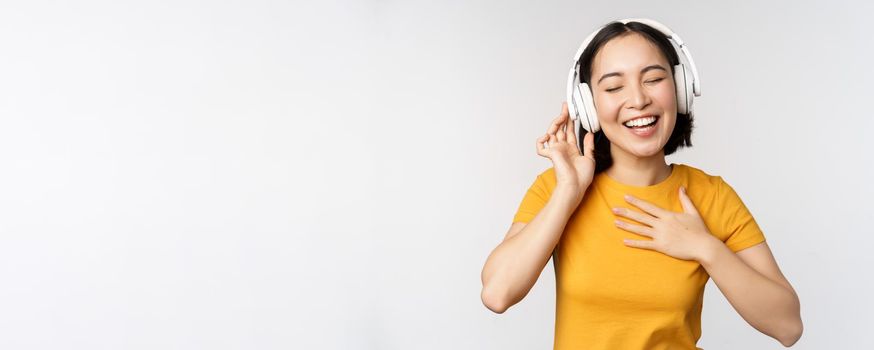 Happy asian girl dancing, listening music on headphones and smiling, standing in yellow tshirt against white background. Copy space
