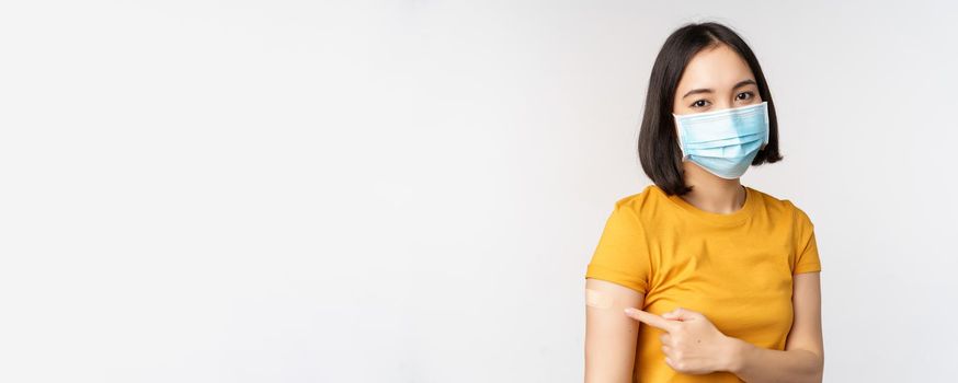 Covid-19, vaccination and healthcare concept. Portrait of cute asian girl in medical mask, has band aid on shoulder after coronavirus vaccine, standing over white background.