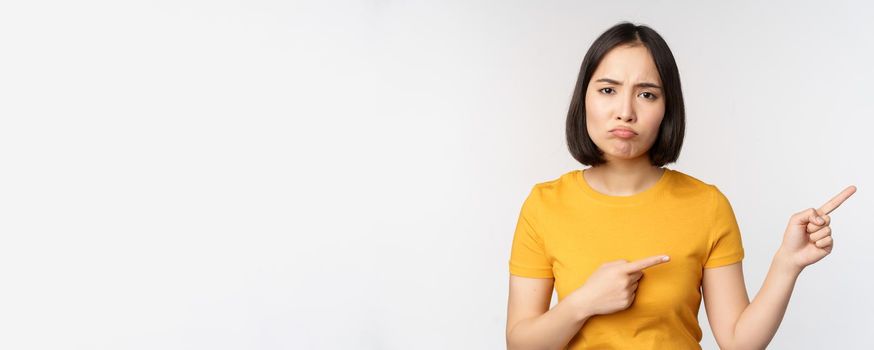 Portrait of disappointed, moody asian girl, pointing fingers right and looking at smth unfair, complaining, standing over white background.