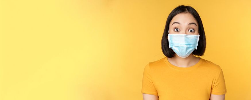 Covid-19 and medical concept. Close up portrait of asian woman in face mask, looking surprised and amazed at news, standing over yellow background.