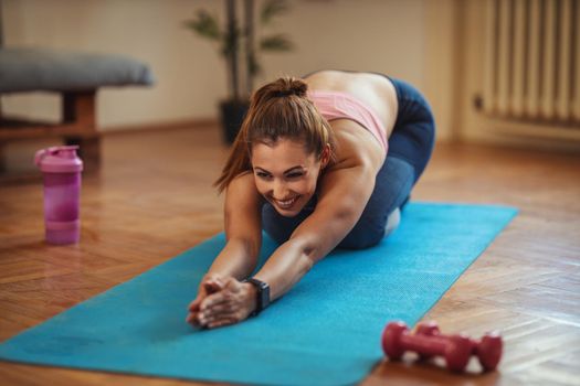 Young smiling woman is doing stretching exercises in the living room on floor mat at home in morning sunshine.