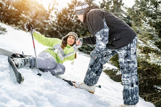 Young man snowboarder helps a beautiful young female skier who fell down on mountain slope.