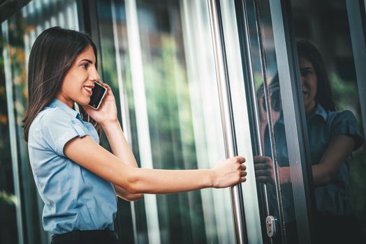 Emotional portrait of a happy and beautiful young businesswoman talking on a smartphone and standing on a business center door.