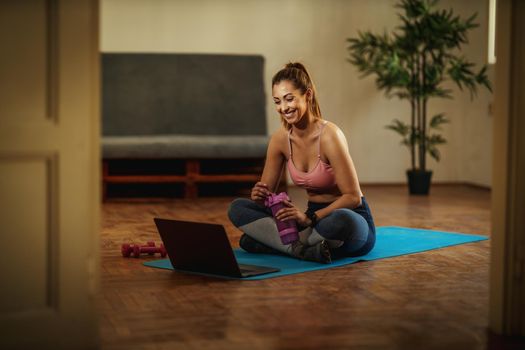 Young smiling woman is watching internet tutorial and doing sports training in the living room on floor mat in front of the laptop.