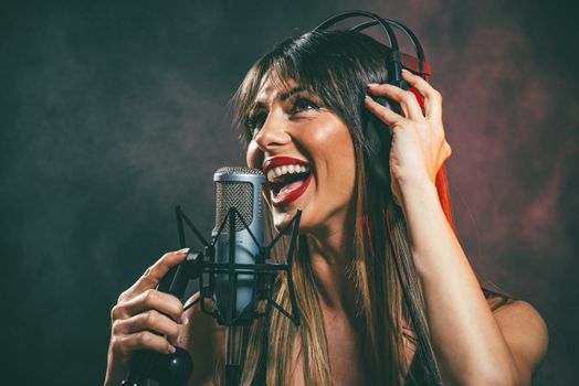 A young woman singer with headphones in front of the microphone. Sing with mouth wide open and with an expression of happiness on her face.