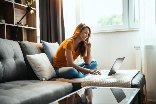 Attractive young woman sitting on the sofa and using her laptop at home.