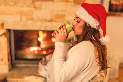 Beautiful young smiling woman enjoying a cup of tea by the fireplace in Christmas evening.