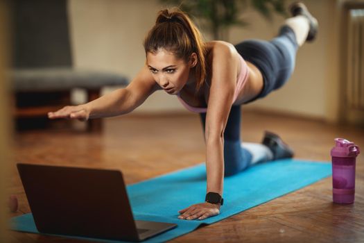 Young smiling woman is doing fitness exercises in the living room on floor mat and looking at the laptop, in morning sunshine.