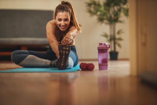 Young smiling woman is doing stretching exercises in the living room on floor mat at home in morning sunshine.